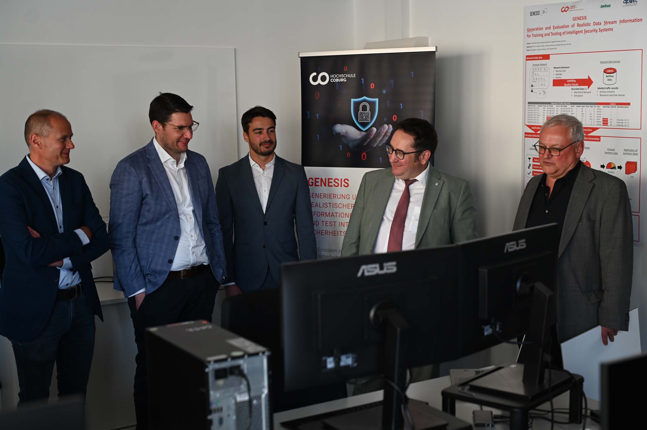 Fünf Männer in Businesskleidung stehen in einem Büro und lächeln auf ASUS-Computermonitore. Plakate und eine Ausstellung zum Thema Cybersicherheit namens „GENESIS“ heben KI-Forschung und CRAI hervor. Im Hintergrund ist auch Staatssekretär Gotthardt zu sehen. at Hochschule Coburg