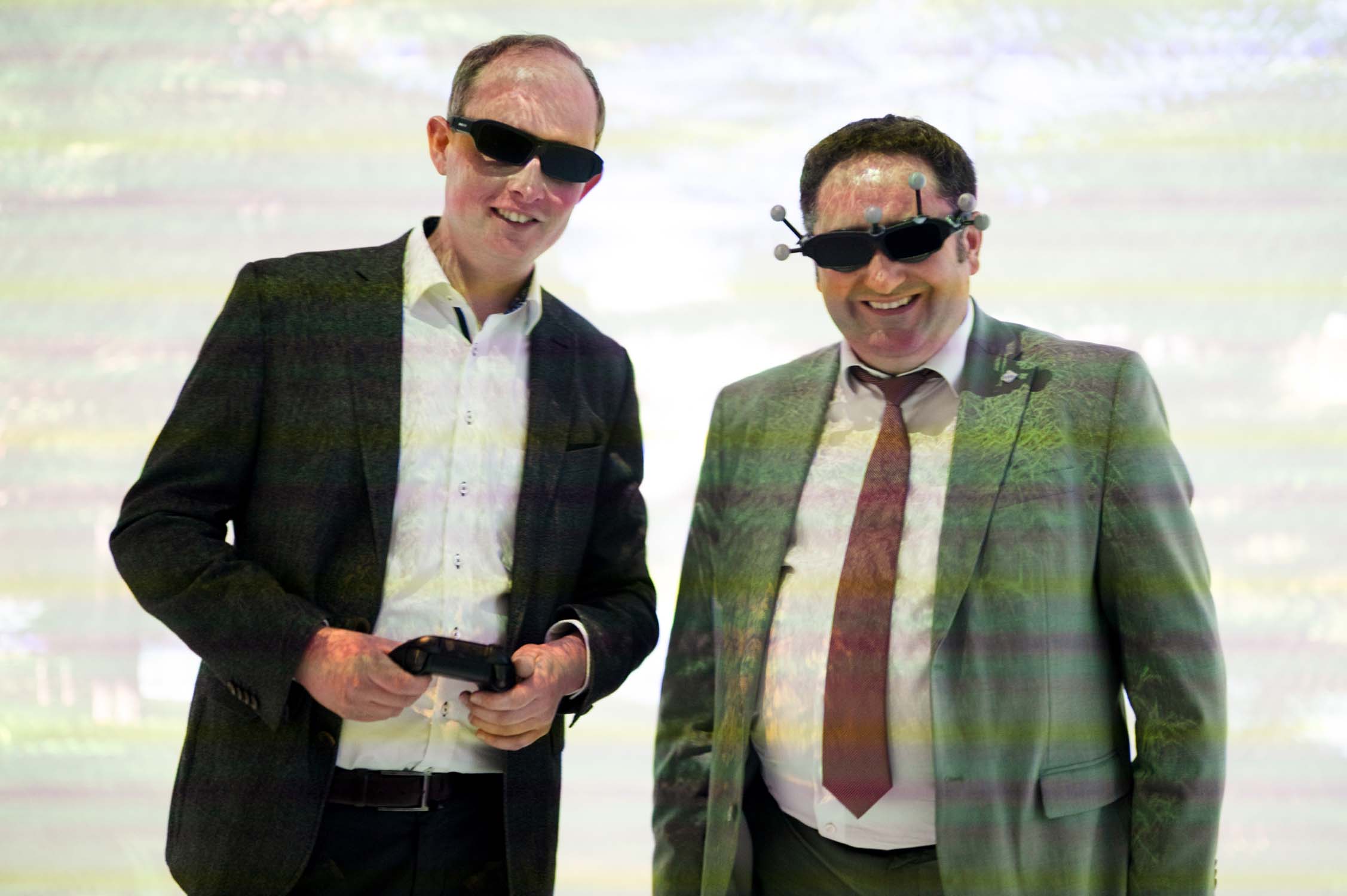 Zwei Männer in Anzügen stehen nebeneinander, beide lächeln und tragen dunkle 3D-Brillen. Staatssekretär Gotthardt hält einen Gamecontroller in der Hand. Ein hell erleuchteter, bunter Hintergrund suggeriert eine digitale oder virtuelle Umgebung, die mit KI-Forschung verbunden ist. at Hochschule Coburg