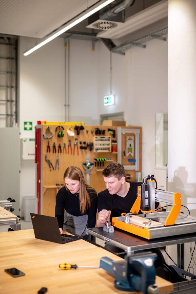 Zwei Personen arbeiten gemeinsam in einer Werkstatt und schauen auf einen Laptop auf einem Holztisch. Im Hintergrund sind Werkzeuge und Geräte zu sehen, daneben steht eine kleine orangefarbene CNC-Maschine. at Hochschule Coburg