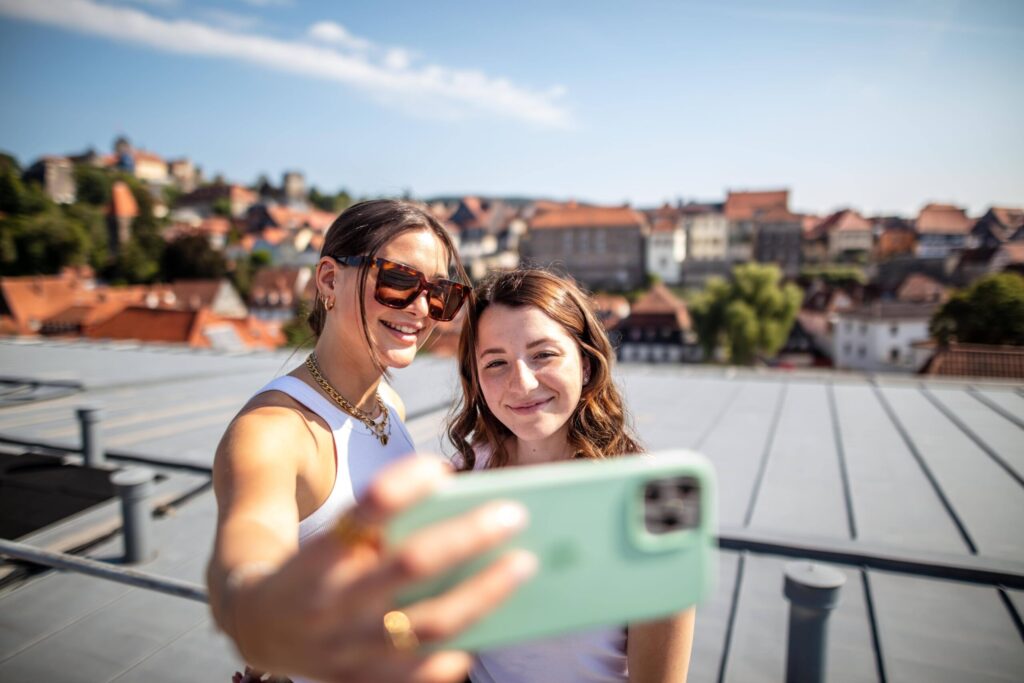 Zwei junge Frauen, die Betriebswirtschaft studieren, lächeln, während sie draußen auf einem Dach ein Selfie machen, mit einem malerischen Blick auf eine Stadt und historische Gebäude im Hintergrund unter einem klaren blauen Himmel. at Hochschule Coburg