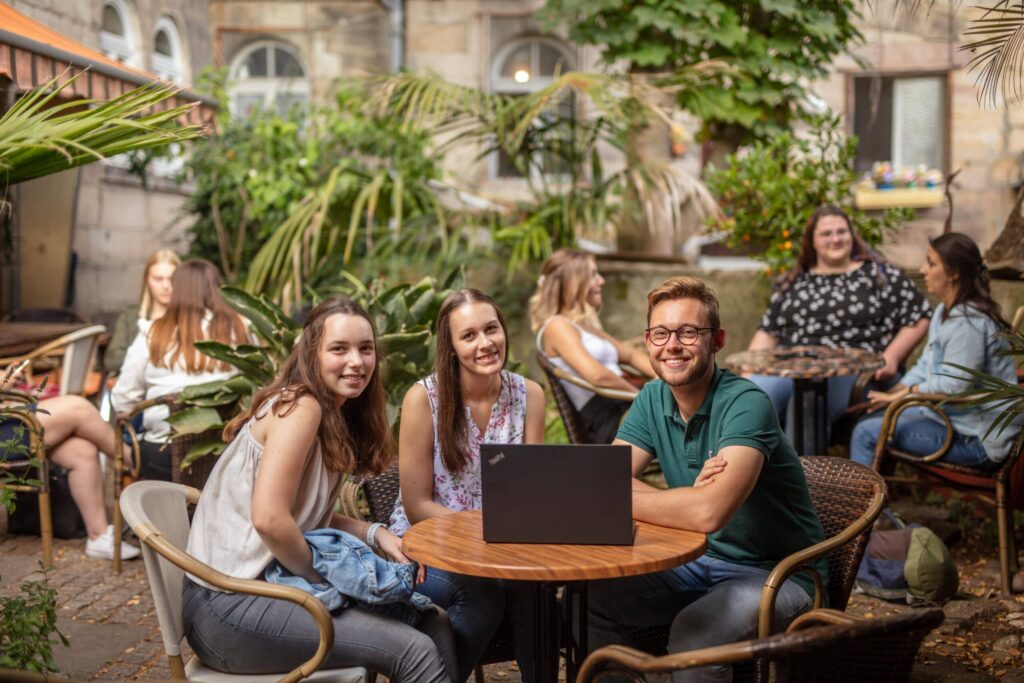 Drei junge Erwachsene sitzen mit einem Laptop an einem Cafétisch im Freien und lächeln in die Kamera – vielleicht diskutieren sie über Betriebswirtschaft. Andere Gruppen unterhalten sich und entspannen an benachbarten Tischen in einem üppigen Garten. at Hochschule Coburg