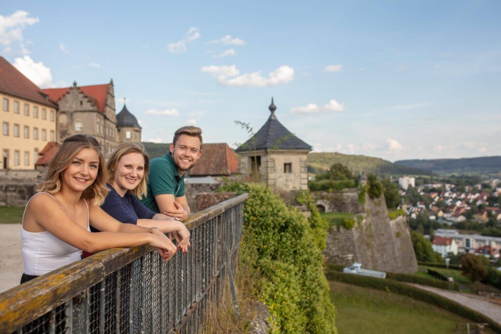 Drei lächelnde Menschen lehnen an einem Metallgeländer mit Blick auf eine malerische, historische Festung – ein inspirierender Ort für Gespräche über Betriebswirtschaft oder Versicherungsmanagement. Im Hintergrund erstrecken sich eine Stadt und sanfte grüne Hügel unter dem blauen Himmel. at Hochschule Coburg