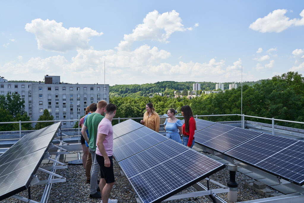 A group of people stand on a rooftop next to solar panels, talking and observing the equipment, with trees and city buildings visible in the background under a partly cloudy sky. at Hochschule Coburg