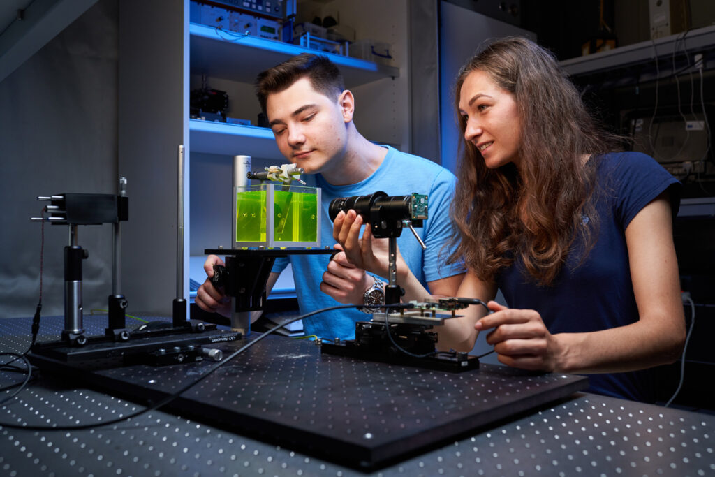 Two young adults in a lab work together using scientific equipment, glass containers, and electronic devices, focusing intently on their engineering experiment as part of their bachelor