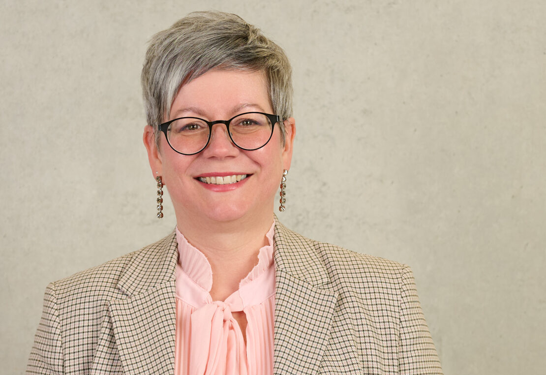 Eine Person mit kurzen grauen Haaren und Brille lächelt. Sie trägt einen hellkarierten Blazer über einer pastellrosa Bluse und lange Ohrringe und steht vor einem schlichten hellen Hintergrund – ein Bild moderner Professorinnen und was eine Frau braucht. at Hochschule Coburg
