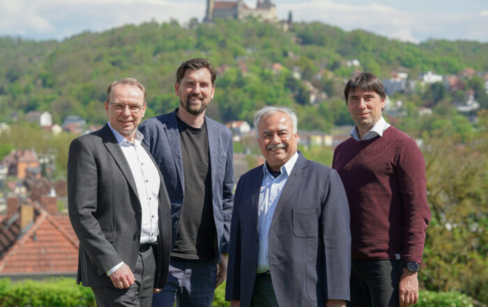 Vier Männer stehen im Freien in legerer Business-Kleidung und lächeln in die Kamera. Im Hintergrund sind ein grüner, bewaldeter Hügel und die Coburger Burg zu sehen. Der klare Himmel und die historische Kulisse lassen vermuten, dass sie diese charmante europäische Stadt besuchen. at Hochschule Coburg