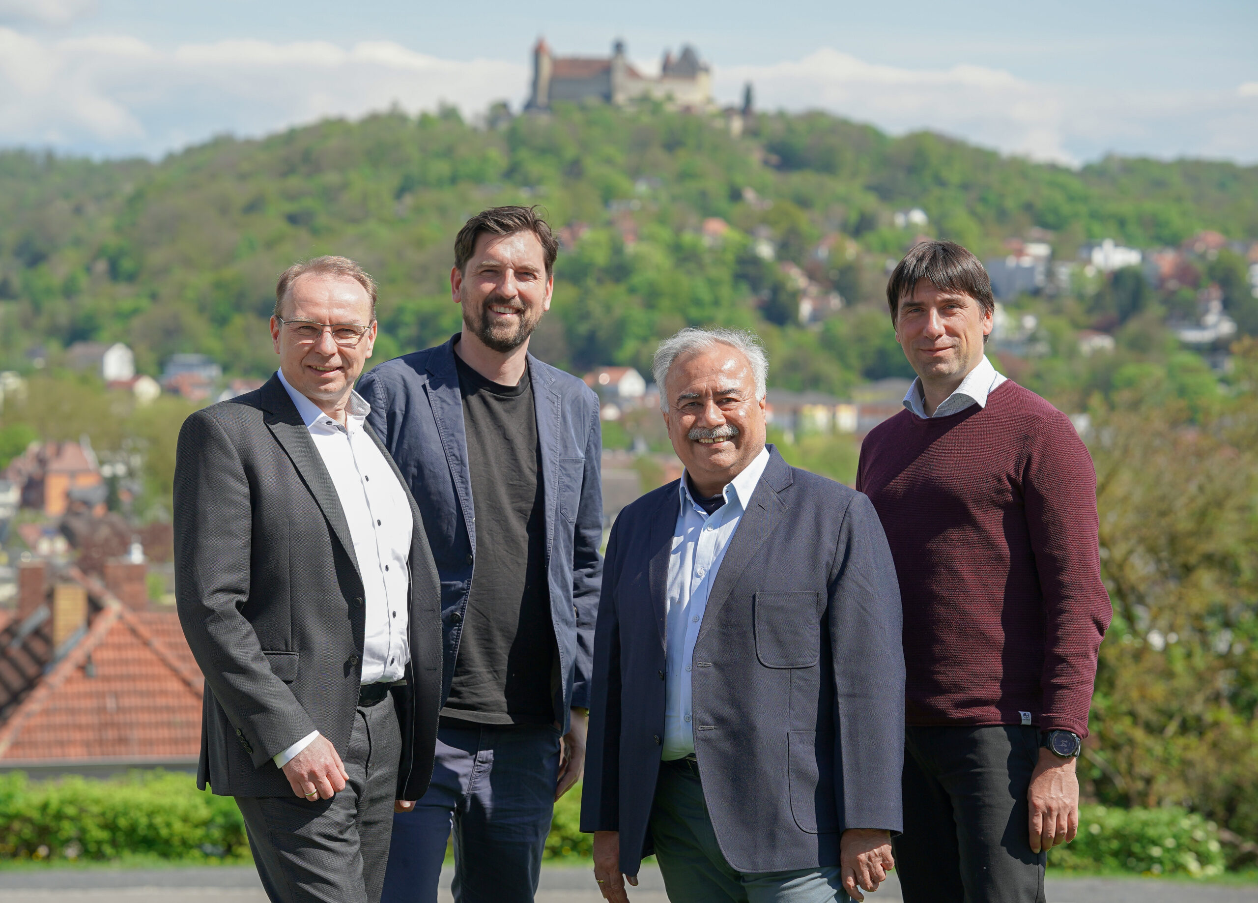 Vier Männer stehen im Freien in legerer Business-Kleidung und lächeln in die Kamera. Im Hintergrund sind ein grüner, bewaldeter Hügel und die Coburger Burg zu sehen. Der klare Himmel und die historische Kulisse lassen vermuten, dass sie diese charmante europäische Stadt besuchen. at Hochschule Coburg