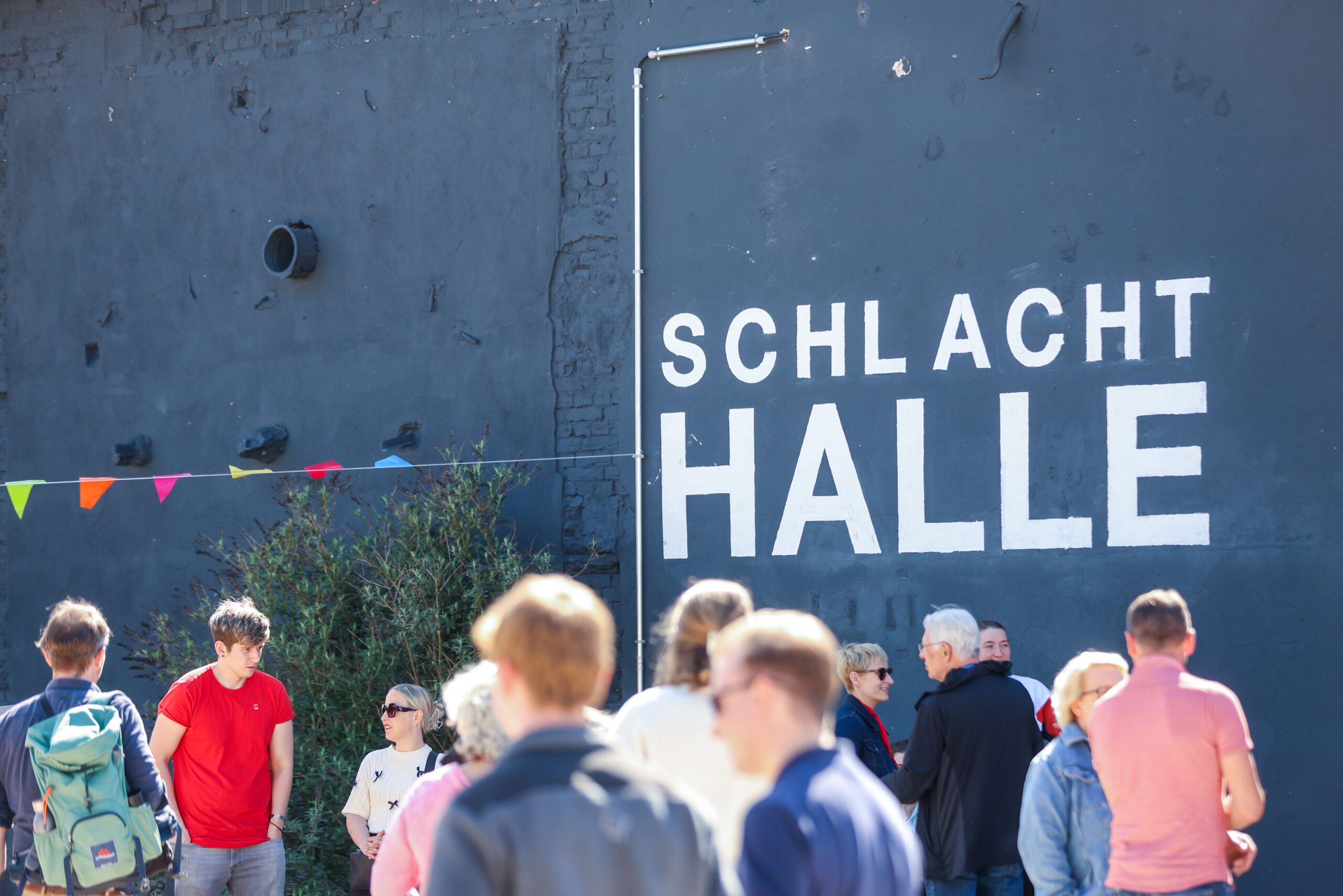 Eine Gruppe von Menschen steht vor dem dunkelgrauen Gebäude der SCHLACHT HALLE und unterhält sich. Bunte Fahnen sorgen für Flair. Die helle, sonnige Atmosphäre lässt auf ein kreatives Treffen der Fakultät Design schließen, vielleicht zur Vorbereitung eines neuen Bühnen- oder Bauprojekts. at Hochschule Coburg