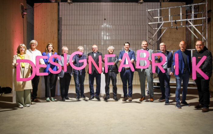 Zwölf Personen stehen im Inneren und halten große rosa Buchstaben mit der Aufschrift „DESIGNFABRIK“. Die Szene im Fakultätsdesign ist von hellen Holzwänden und weißen Fliesen geprägt, während auf der rechten Seite ein Metallgerüst an eine bühnenartige Bühne erinnert. at Hochschule Coburg