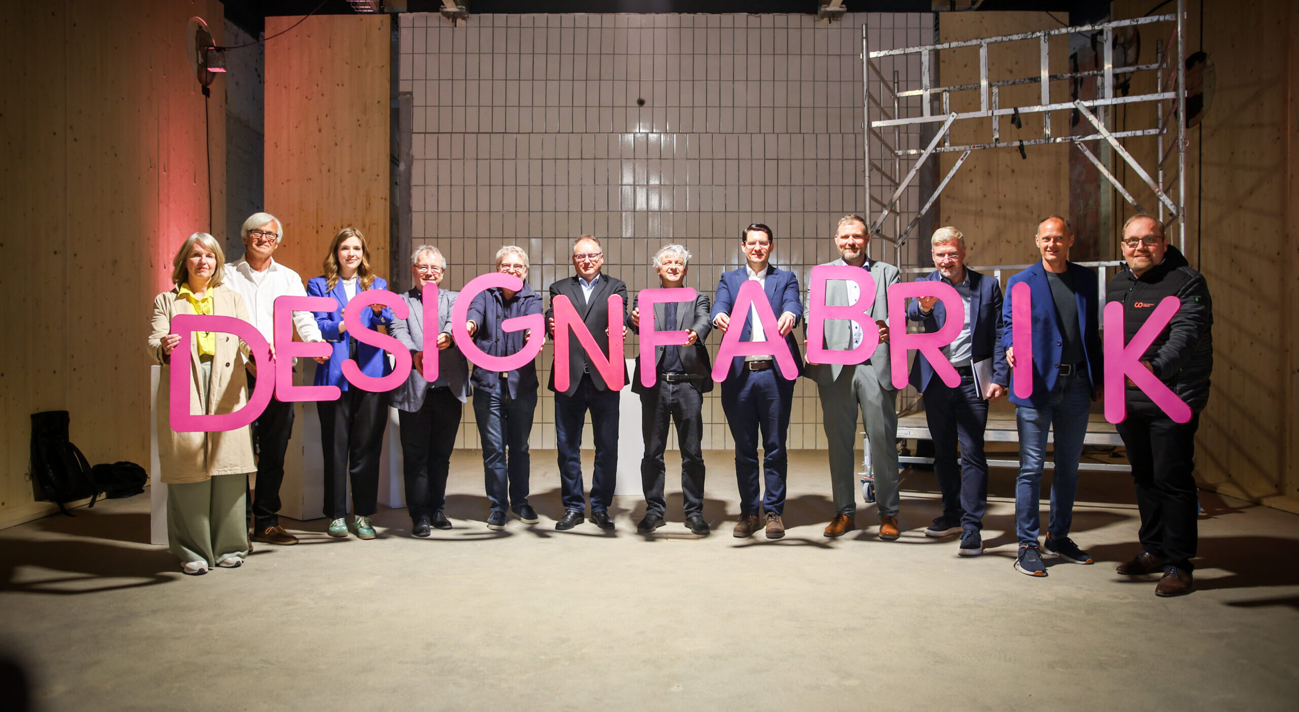 Zwölf Personen stehen im Inneren und halten große rosa Buchstaben mit der Aufschrift „DESIGNFABRIK“. Die Szene im Fakultätsdesign ist von hellen Holzwänden und weißen Fliesen geprägt, während auf der rechten Seite ein Metallgerüst an eine bühnenartige Bühne erinnert. at Hochschule Coburg