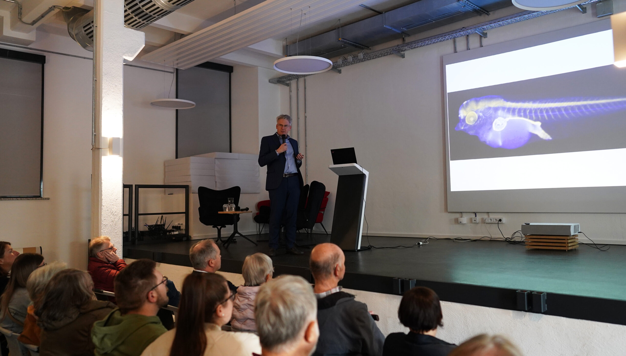 Ein Mann im Anzug hält in einem modernen Raum vor einem Publikum einen Vortrag, bei dem er „Wissen für alle“ vermittelt und dabei auf einer großen Leinwand das farbenfrohe wissenschaftliche Bild eines Fisches zeigt. at Hochschule Coburg