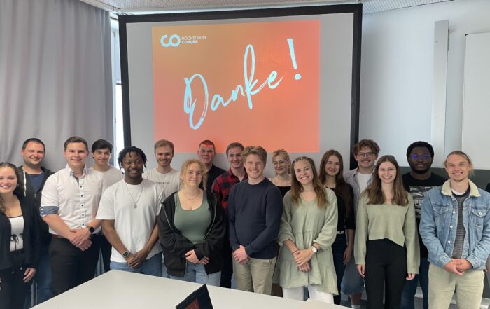 Eine Gruppe von 17 jungen Erwachsenen, die Mentorinnen und Mentoren, steht lächelnd in einem Klassenzimmer vor einem Bildschirm, auf dem in großen Buchstaben „Danke!“ auf rotem Grund steht. Auf dem Tisch im Vordergrund sind ein Laptop und andere Gegenstände zu sehen. at Hochschule Coburg