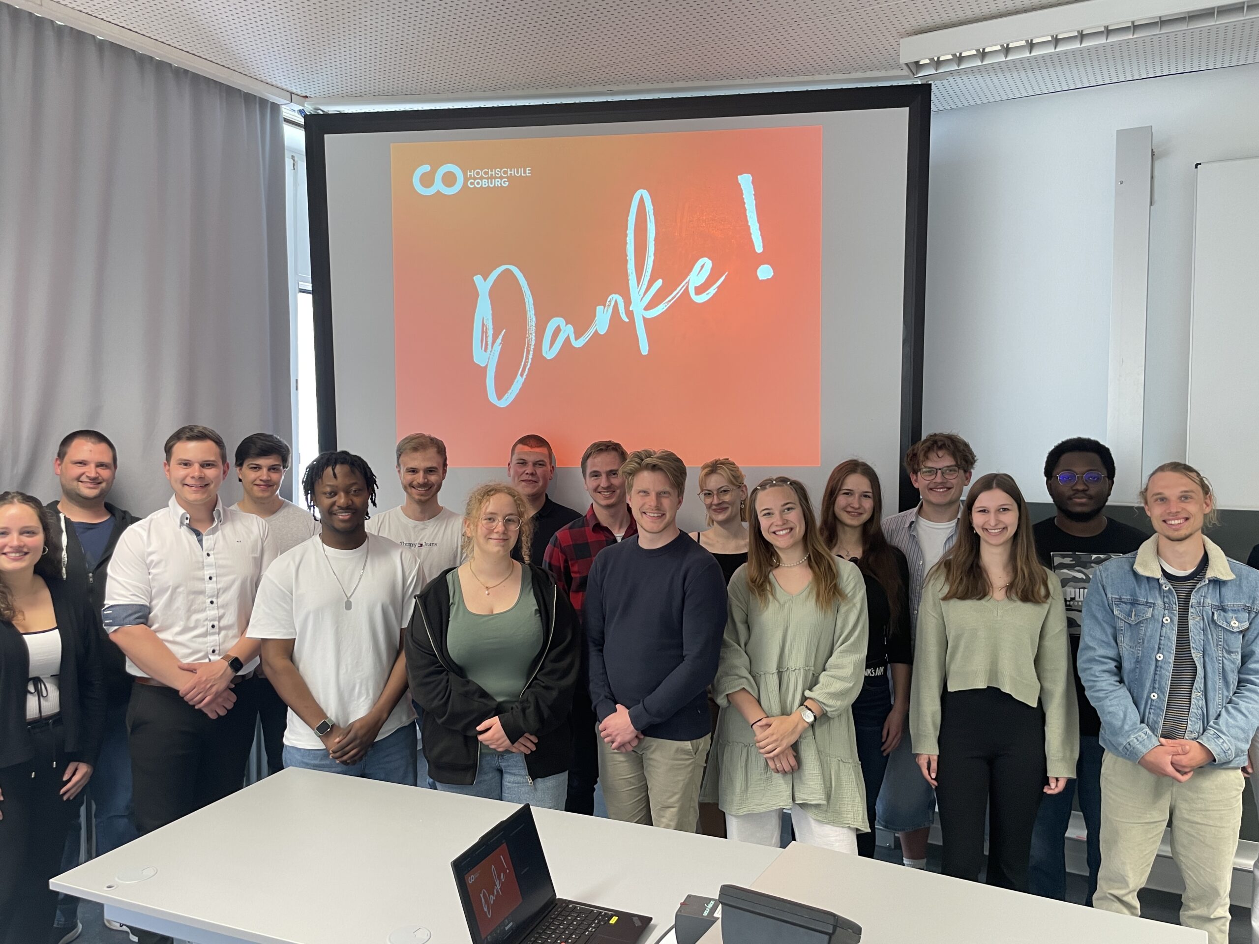 Eine Gruppe von 17 jungen Erwachsenen, die Mentorinnen und Mentoren, steht lächelnd in einem Klassenzimmer vor einem Bildschirm, auf dem in großen Buchstaben „Danke!“ auf rotem Grund steht. Auf dem Tisch im Vordergrund sind ein Laptop und andere Gegenstände zu sehen. at Hochschule Coburg