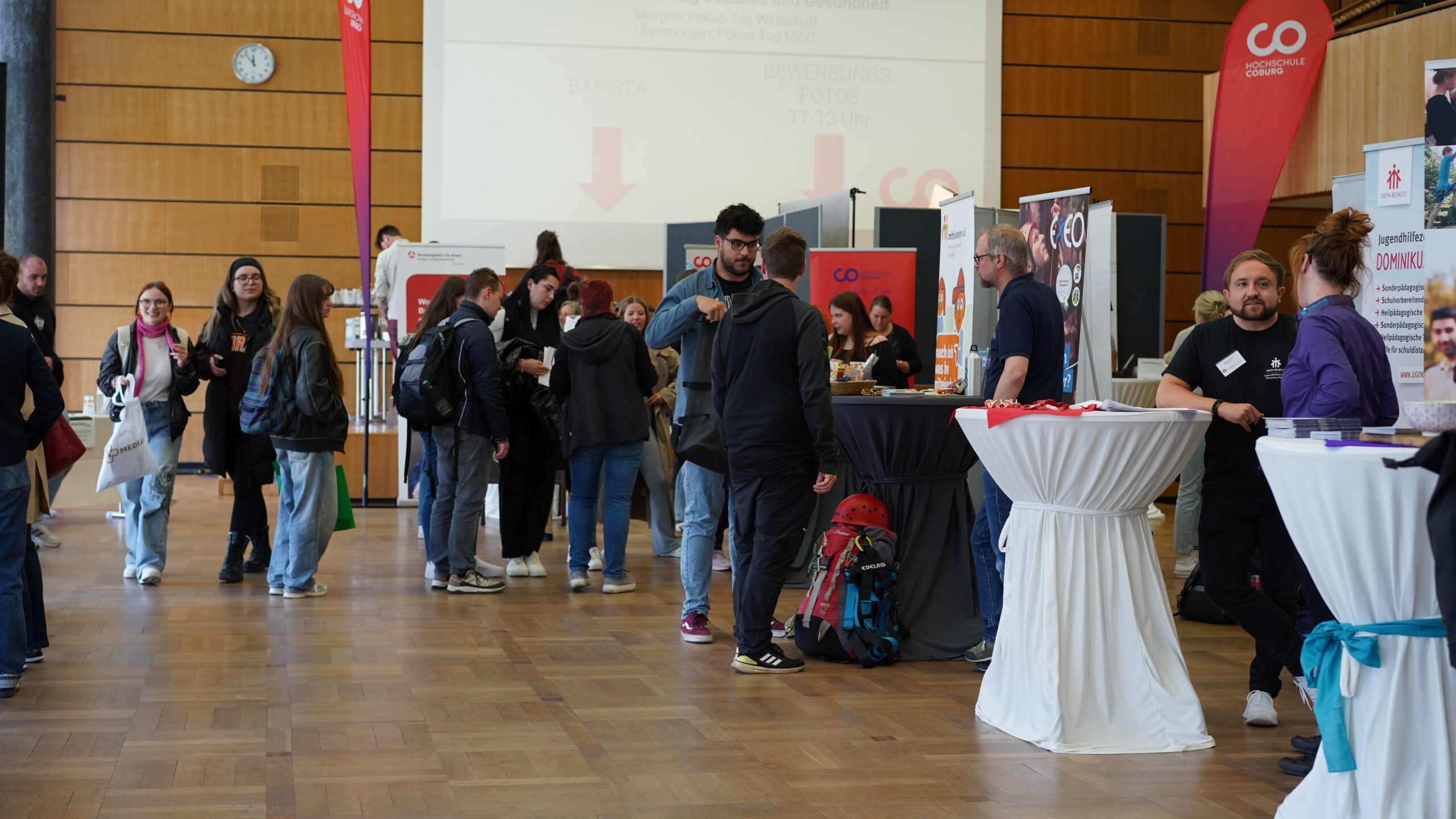Menschen treffen sich bei einer Indoor-Veranstaltung mit Informationsständen und Bannern und tauschen sich aus. Manche stehen an Stehtischen, andere gehen oder unterhalten sich in Gruppen. Die Atmosphäre ist locker und lebendig, mit holzgetäfelten Wänden im Hintergrund. at Hochschule Coburg
