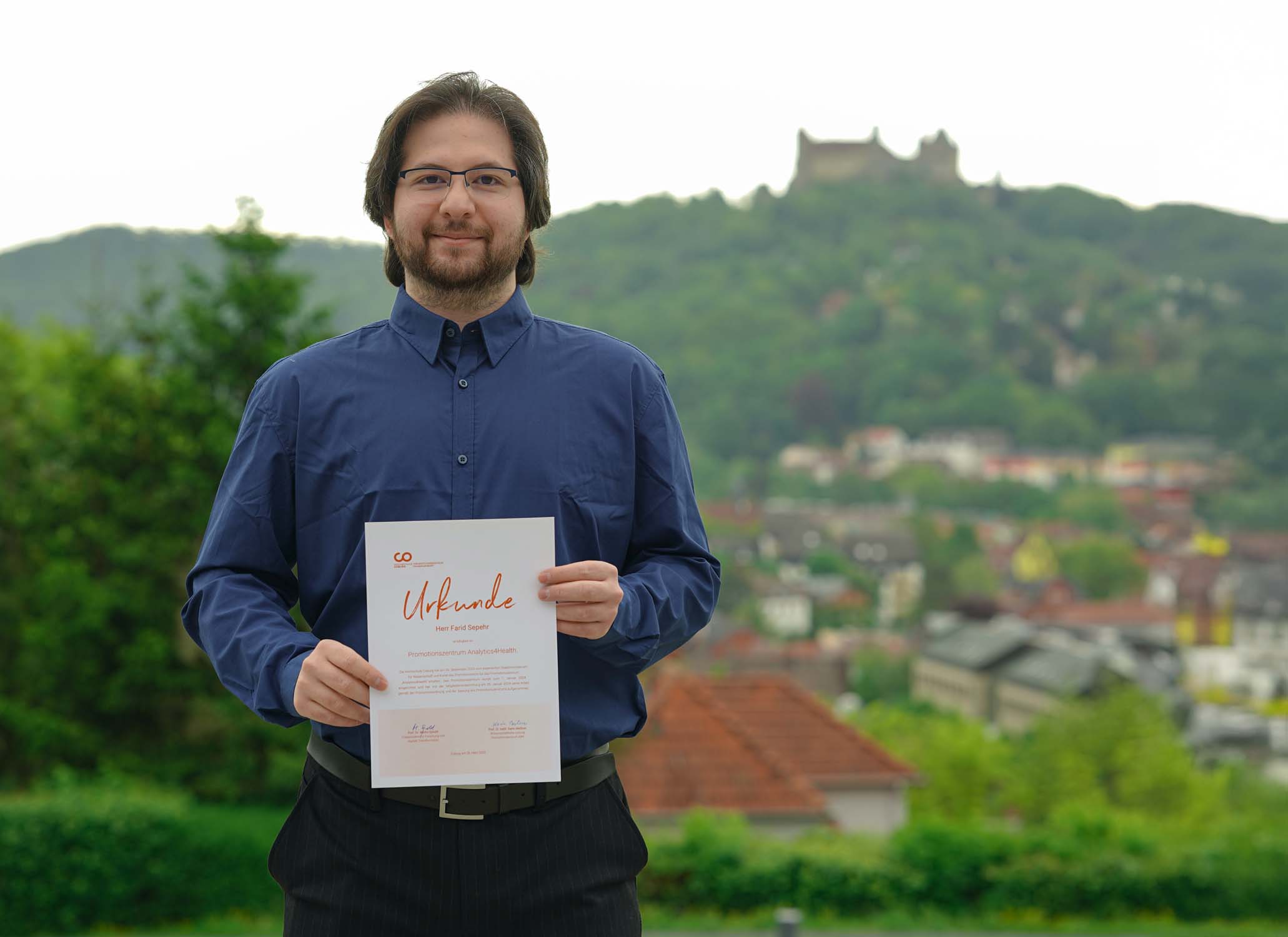 Ein Mann mit Brille und blauem Hemd steht lächelnd im Freien und hält eine Urkunde in der Hand. Im Hintergrund ist eine malerische Aussicht auf eine Stadt und ein Schloss auf einem grünen Hügel zu sehen – er feiert seinen Doktortitel im Bereich Klimaschutz. at Hochschule Coburg