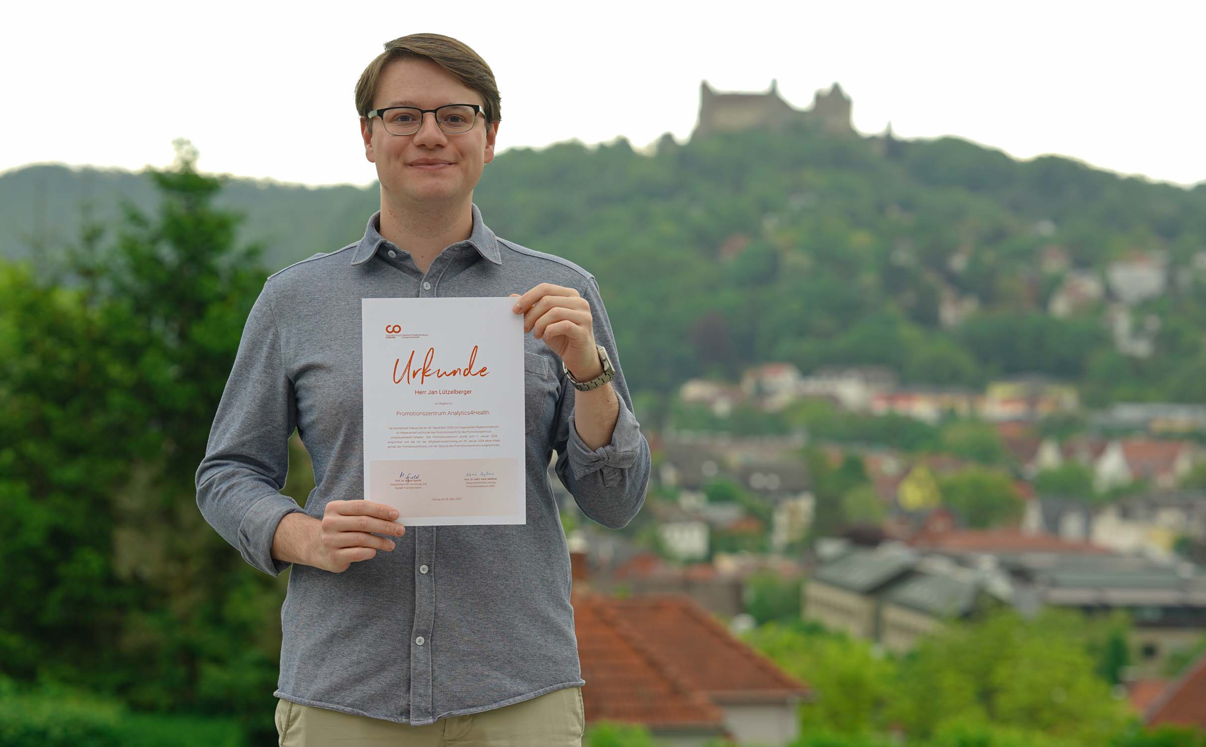 Ein Mann mit Brille steht im Freien, hält ein Zertifikat in der Hand und lächelt stolz. In der grünen Hügellandschaft hinter ihm thront eine Burg über einer Stadt – vielleicht feiert er gerade seinen Doktortitel im Bereich Klimaschutz oder KI. at Hochschule Coburg