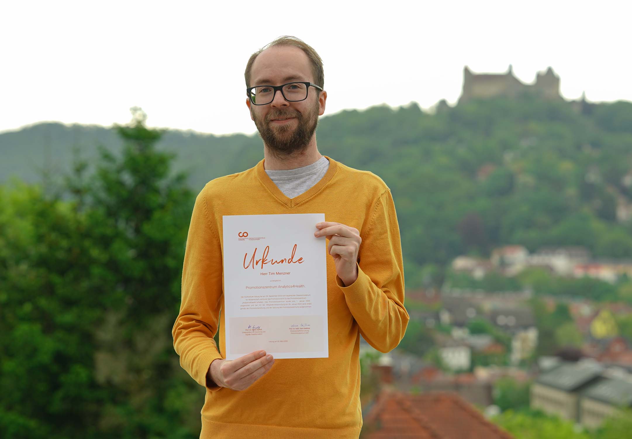 Ein Mann mit Brille und gelbem Pullover hält im Freien eine Urkunde für seinen Doktortitel hoch, im Hintergrund sind Hügel, Bäume und ein Schloss verschwommen zu sehen. at Hochschule Coburg