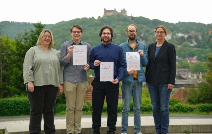 Fünf Personen stehen im Freien, drei halten Zertifikate in den Händen und lächeln. Mit einer Burg auf einem Hügel im Hintergrund wirkt die Gruppe glücklich und lässig gekleidet. Möglicherweise feiert sie einen Erfolg im Klimaschutz oder den Abschluss eines Doktortitels. at Hochschule Coburg