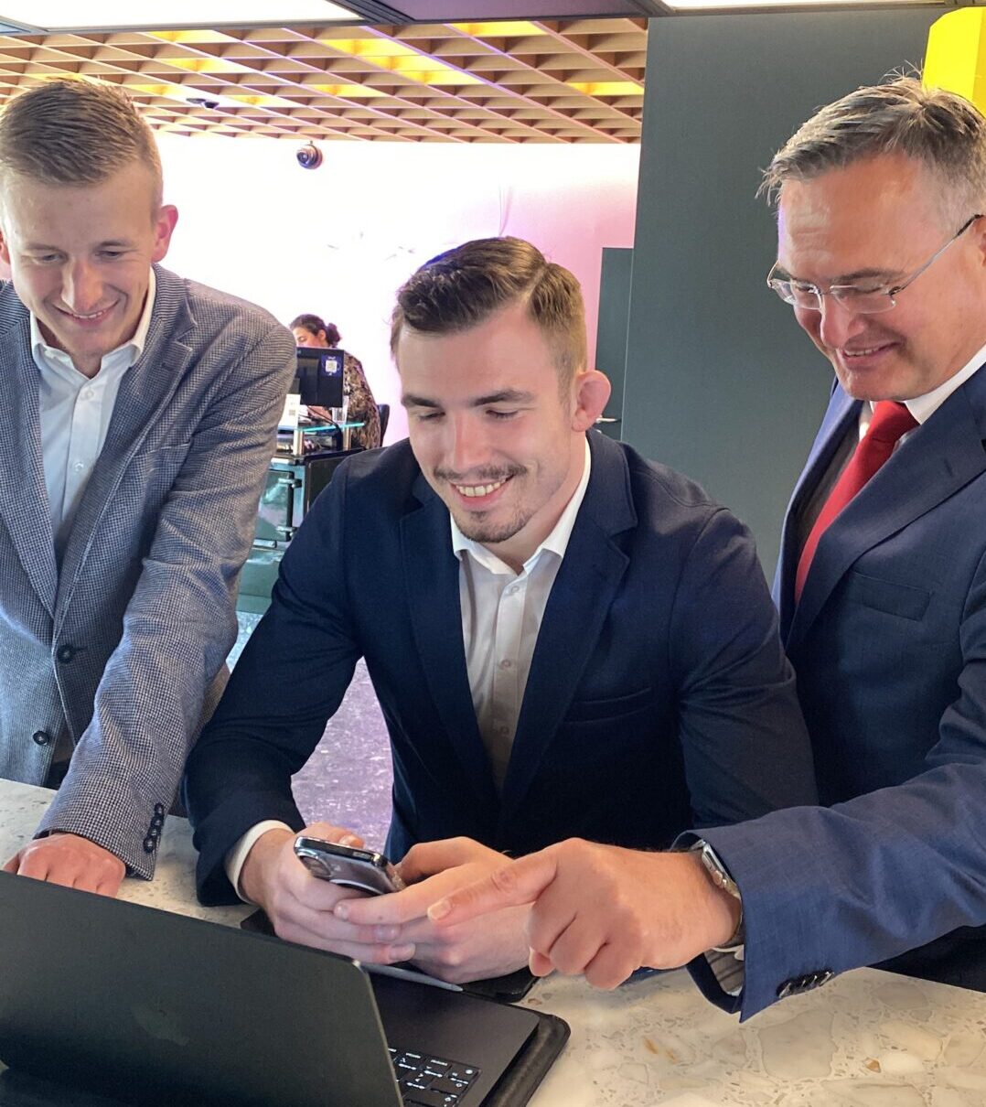 Drei Männer in Anzügen stehen lächelnd an einem Tresen und schauen auf einen Laptop. Einer hält ein Smartphone in der Hand, die anderen schauen zu, was auf Teamarbeit hindeutet – vielleicht für ein deutsch-bulgarisches Studien-Projekt und grenzenloses Wissen. at Hochschule Coburg