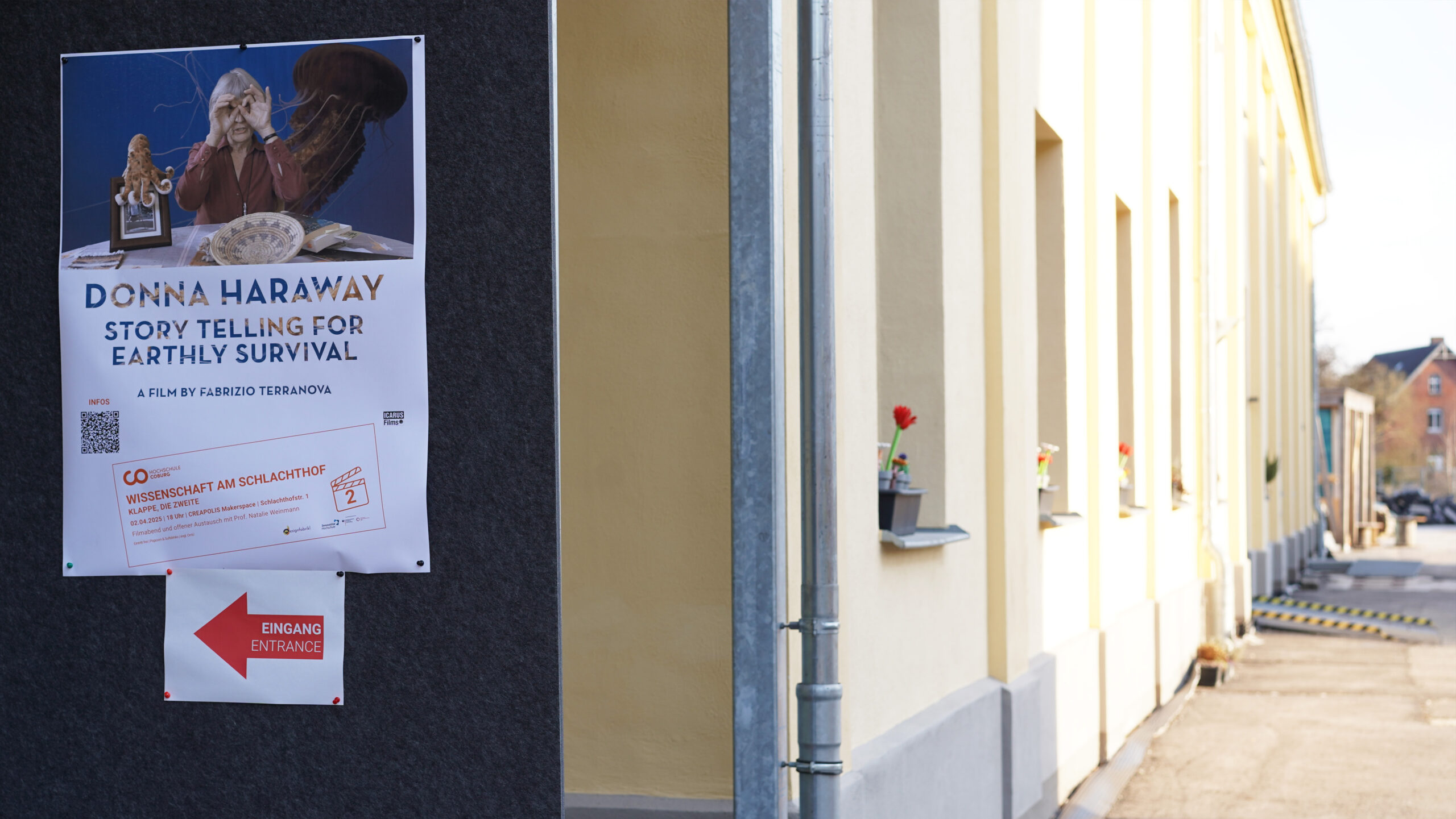 Ein Plakat zum Film „Donna Haraway: Geschichten erzählen fürs Überleben“ prangt an einer dunklen Wand vor einem gelben Gebäude, das an einen Schlachthof erinnert. Ein roter Pfeil weist nach links zum Eingang; Blumen erhellen stille Fensterbänke. at Hochschule Coburg
