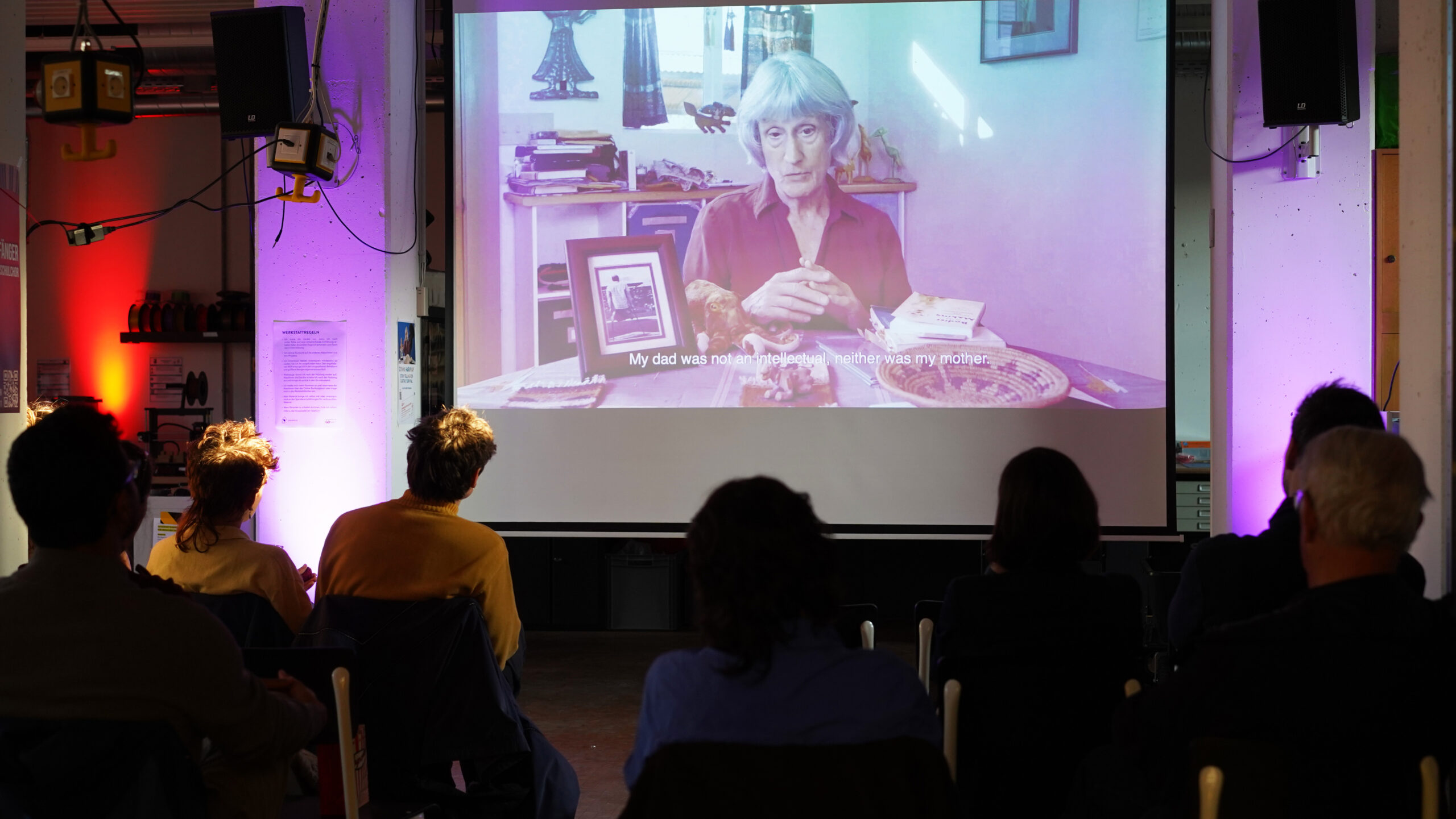 Eine Gruppe von Menschen sitzt in einem dunklen Raum und sieht sich einen auf eine Leinwand projizierten Film an. Zu sehen ist Donna Haraway – eine grauhaarige Frau –, die an einem Tisch spricht, auf dem Fotos, Bücher und Untertitel zu sehen sind. at Hochschule Coburg