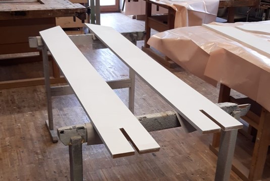 Zwei lange, weiß lackierte Holzplatten liegen auf Metallgestellen in einer Werkstatt der Hochschule Coburg. An einem Ende ist jeweils eine rechteckige Aussparung eingearbeitet. Im Hintergrund sind Arbeitstische und Werkzeuge zu sehen. at Hochschule Coburg