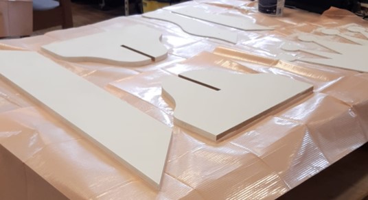 Mehrere flache, weiße Holzteile mit Schlitzen und Rundungen liegen auf einem mit einer Plastikfolie abgedeckten Tisch in der Hochschule Coburg, vermutlich vorbereitet für den Zusammenbau oder die Montage. at Hochschule Coburg