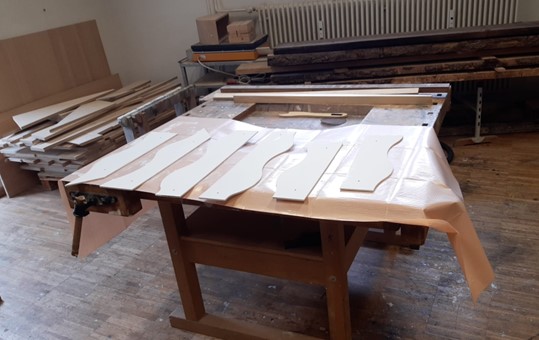 Auf einer Holzwerkbank in einer Werkstatt, vermutlich der Hochschule Coburg, liegen sechs weiße, gebogene Holzteile. Im Hintergrund sind verschiedene Werkzeuge und Bauholz zu sehen. Die Oberfläche ist mit Plastikfolie abgedeckt. at Hochschule Coburg