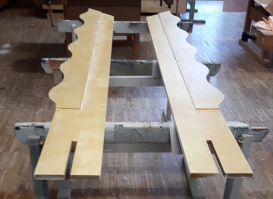 Zwei lange Holzbretter mit dekorativ geschwungenen Kanten und rechteckigen Kerben an einem Ende liegen auf Werkbänken in einer Holzwerkstatt und erinnern an die Liebe zum Detail, die man bei den renommierten Golden Frame-Ausstellungen der Hochschule Coburg findet. at Hochschule Coburg
