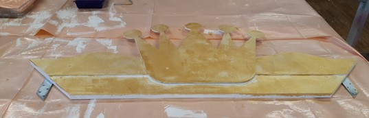 Ein großer, goldbemalter Kronenausschnitt aus Holz steht auf einem kunststoffgedeckten Tisch im Café Victoria. Die Krone hat fünf abgerundete Spitzen, flankiert von eckigen Formen und Metallklammern an beiden Enden und ähnelt damit einem goldenen Rahmen der Hochschule Coburg. at Hochschule Coburg