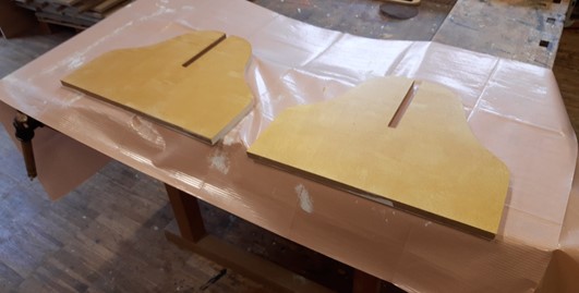 Zwei gelbe, symmetrische Holzbretter mit mittigen Schlitzen liegen auf einer kunststoffbeschichteten Werkbank in einer Werkstatt der Hochschule Coburg, umgeben von Sägemehl und Holzoberflächen. at Hochschule Coburg