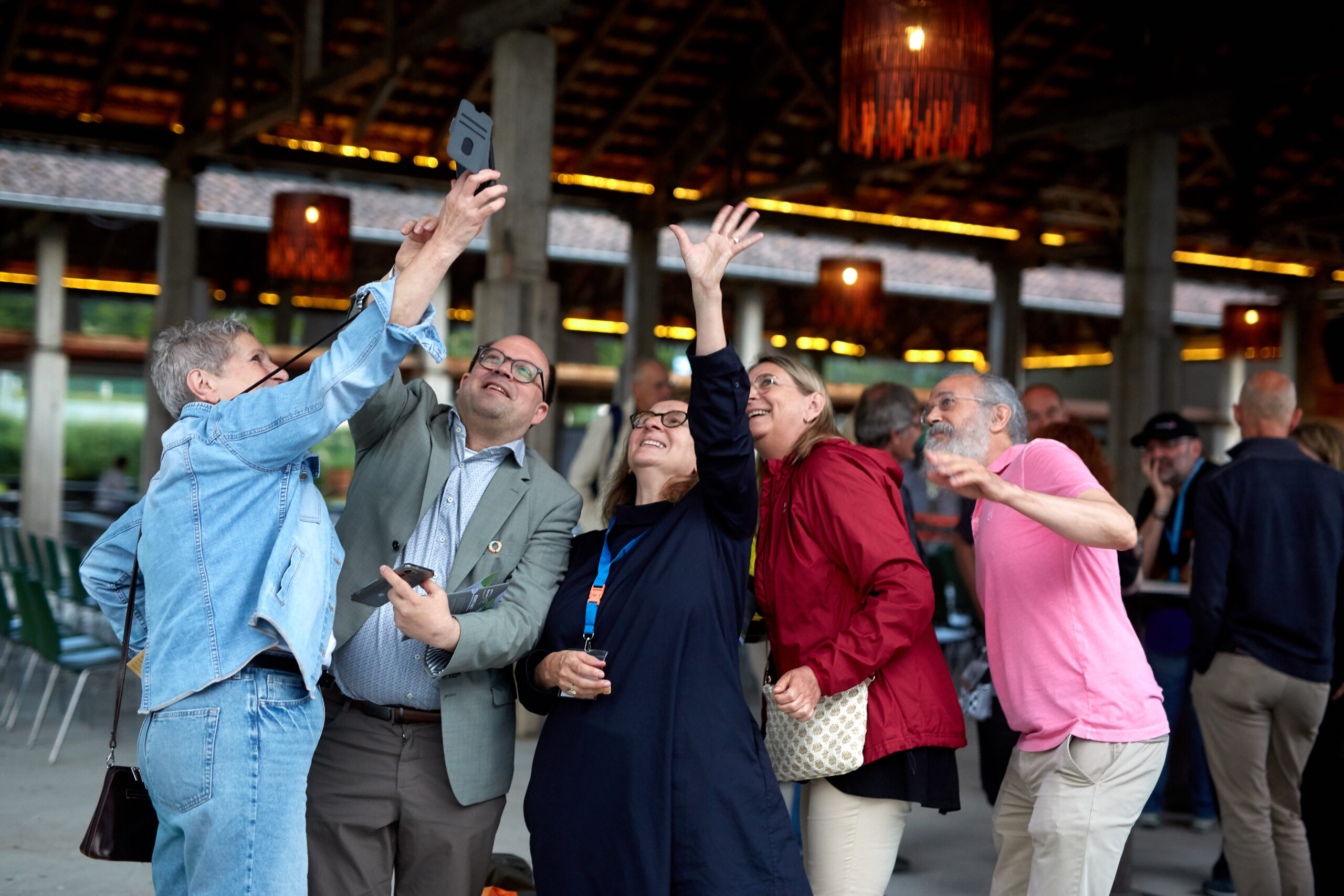 Coburger_Nachhaltigkeitstage_2024_71 Eine Gruppe von sechs Erwachsenen steht dicht beieinander unter einem Pavillon in Coburg, lächelt und winkt, während sie während der lebhaften Coburger Nachhaltigkeitstage ein Selfie machen, wobei einige Telefone halten und andere aufgeregt gestikulieren. at Hochschule Coburg