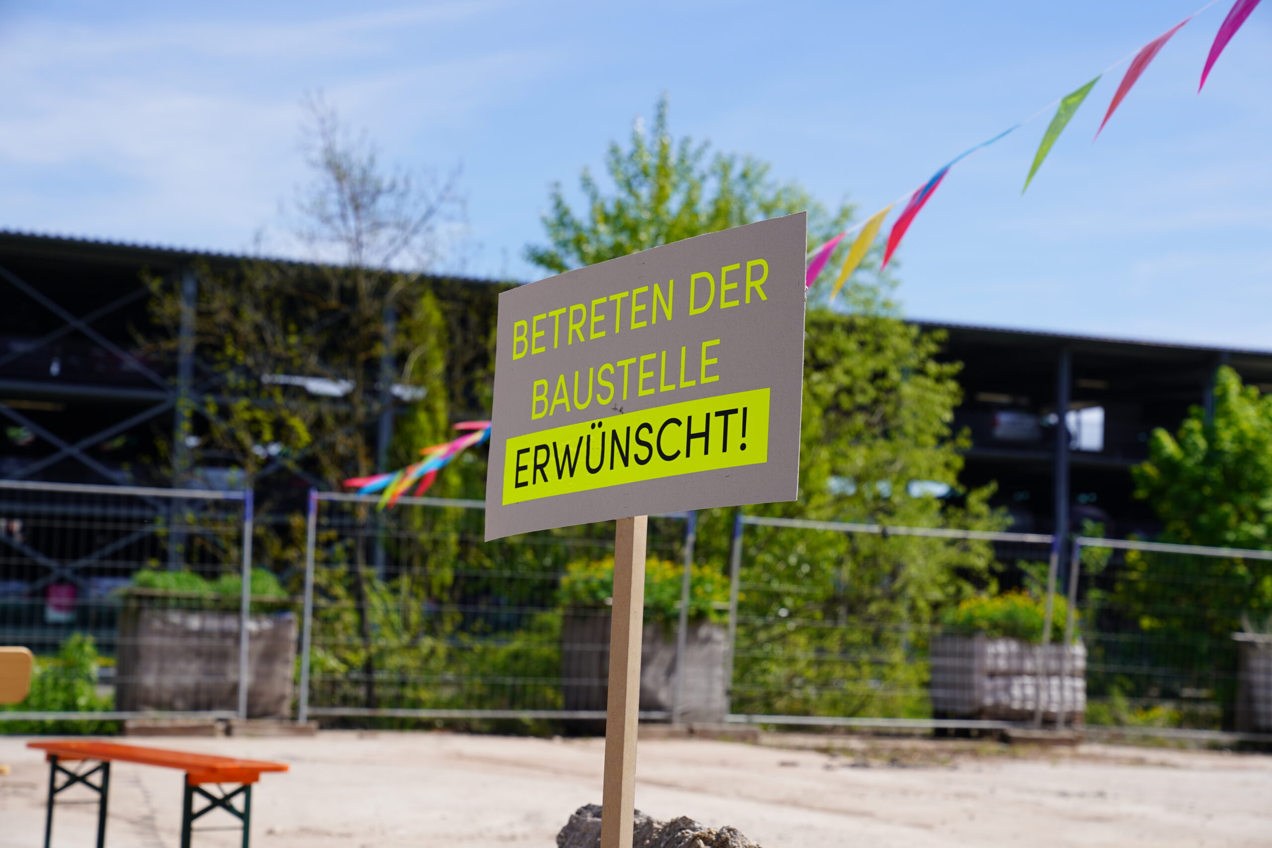 Auf einer Baustelle steht ein Schild mit der Aufschrift „Betreten der Baustelle erwünscht!“. Bunte Wimpel und Bäume rahmen diese lebendige Bühne für Kreativität ein, passend zur Fakultät Design. at Hochschule Coburg