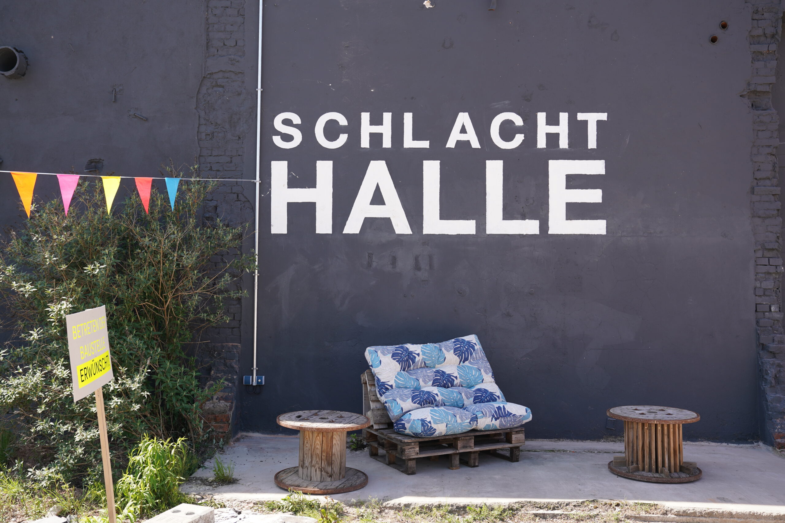 Vor einer dunkelgrauen Wand steht ein gepolstertes Sofa aus Holzpaletten, auf dem in großen weißen Buchstaben „SCHLACHT HALLE“ steht. In der Nähe hängen bunte Wimpel, davor steht auf dem Boden ein gelbes Schild der Fakultät Design. at Hochschule Coburg