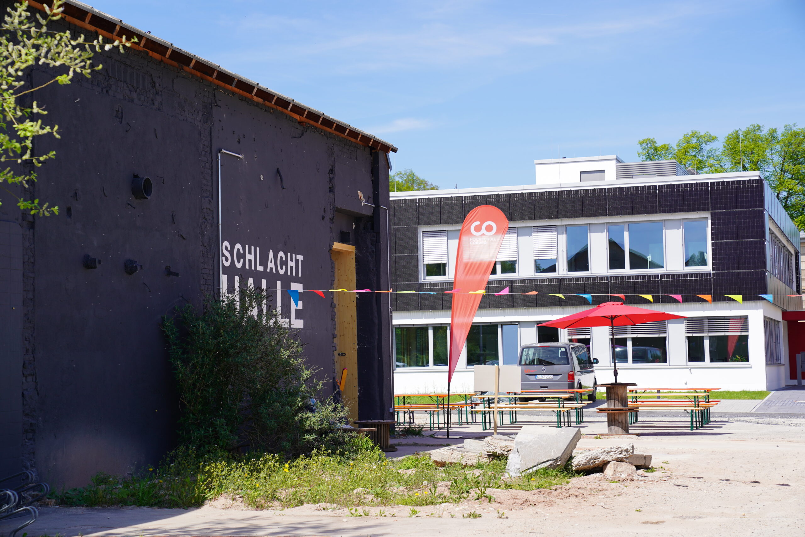 Ein schwarzes Gebäude mit der Aufschrift „Schlacht Hall“ an der Wand steht neben einem modernen weißen Gebäude – dem Sitz der Fakultät Design. Eine rote Fahne, bunte Wimpel und Tische im Freien mit roten Sonnenschirmen sorgen an einem sonnigen Tag für eine lebendige Bühne. at Hochschule Coburg