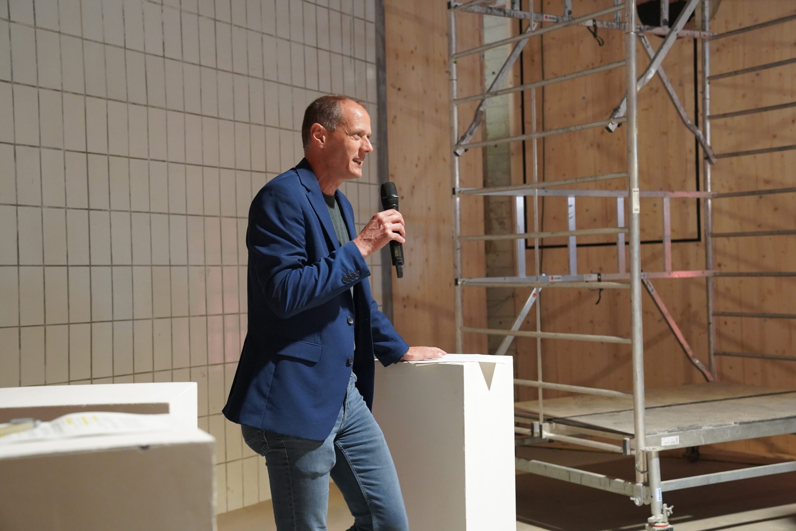 Ein Mann in blauem Blazer und Jeans spricht in ein Mikrofon auf der Bühne, neben einem weißen Podium. Hinter ihm befinden sich geflieste und Holzwände, Metallgerüste und Elemente, die an Bauen erinnern und den kreativen Geist der Fakultät Design widerspiegeln. at Hochschule Coburg