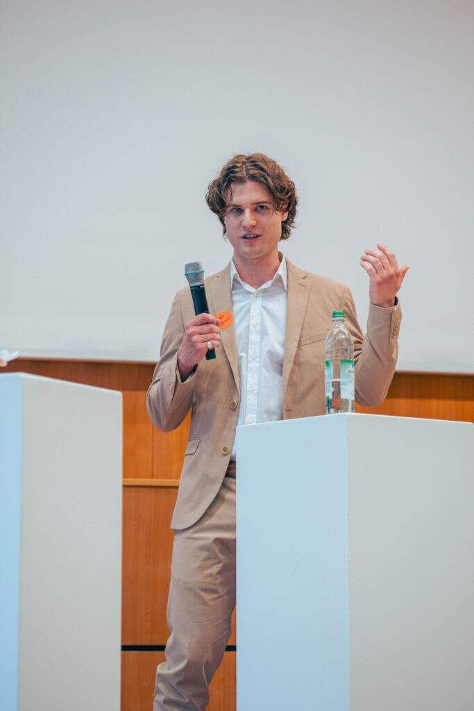 Ein junger Mann in einem beigen Anzug spricht in ein Mikrofon, während er hinter einem weißen Podium mit einer Wasserflasche darauf steht, in einem Innenbereich vor einem hölzernen und weißen Hintergrund. at Hochschule Coburg
