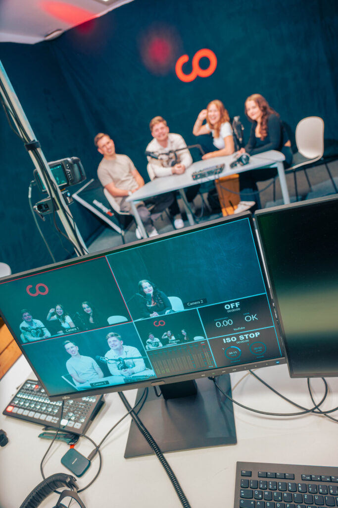 Auf einem Computermonitor wird ein Live-Video von vier jungen Leuten angezeigt, die in einem Studio an einem Tisch mit Mikrofonen sitzen, während im Hintergrund die Gruppe zu sehen ist, wie sie sitzt und lächelt. at Hochschule Coburg
