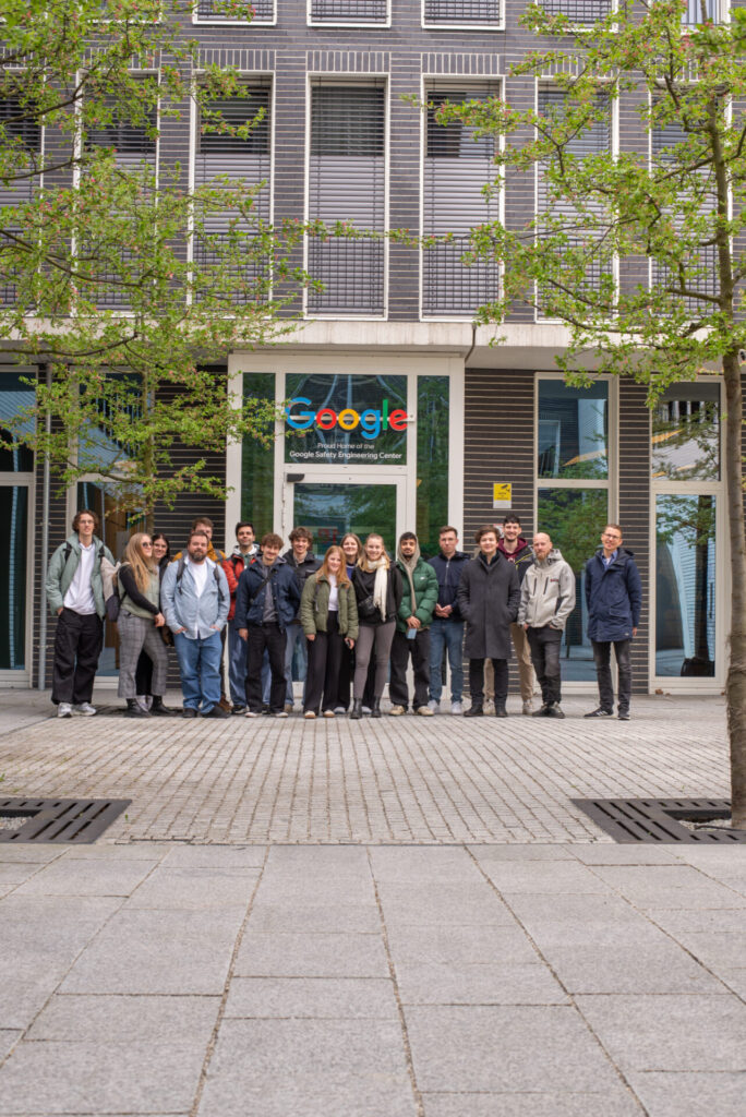 A group of people standing and smiling in front of a Google office building, framed by green trees and a paved courtyard. at Hochschule Coburg