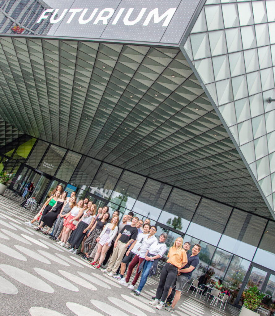 A group of people stand in a line outside the Futurium building, which features a modern glass facade and a geometric patterned entrance. The sign "Futurium" is prominently displayed above them. at Hochschule Coburg
