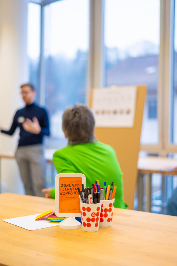 Das Bild konzentriert sich auf einen Tisch mit Stiften und einem Schild mit der Aufschrift „Zukunft Lernen Workshop“ und zeigt zwei Personen im Gespräch neben einem Flipchart in einem hellen, modernen Raum. at Hochschule Coburg