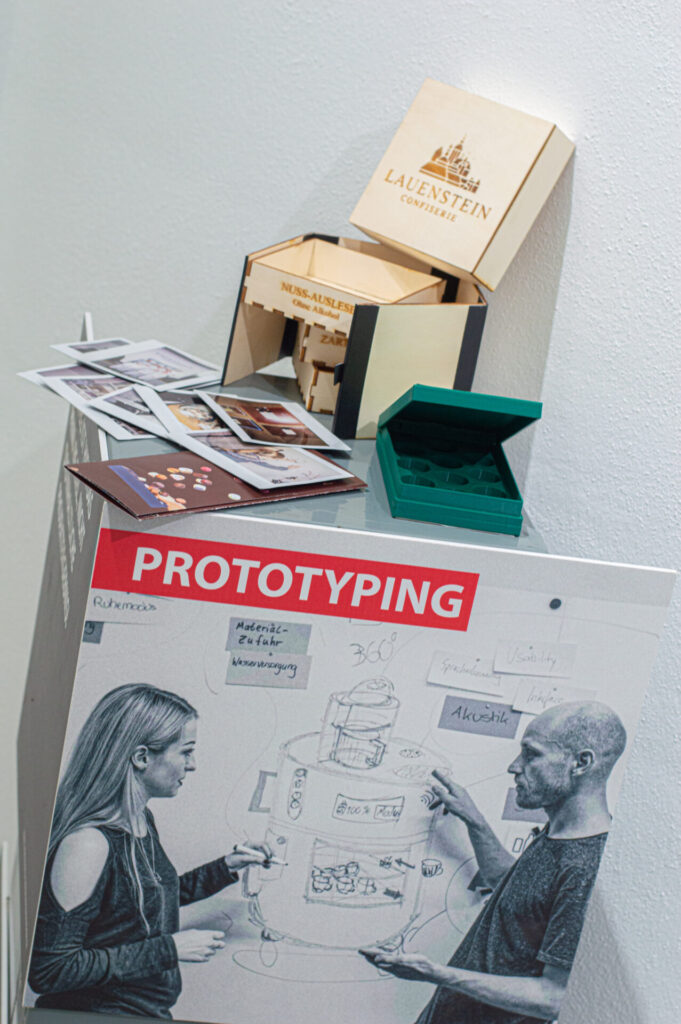 Ein Display mit einem „PROTOTYPING“-Poster, das zwei Personen zeigt, die einen Entwurf besprechen, mehreren Fotos, einer grünen Box und einer darauf angeordneten Holzbox „Lauenstein Confiserie“ – perfekt für die Präsentation von Ideen in der Wirtschaftsinformatik 2.0. at Hochschule Coburg