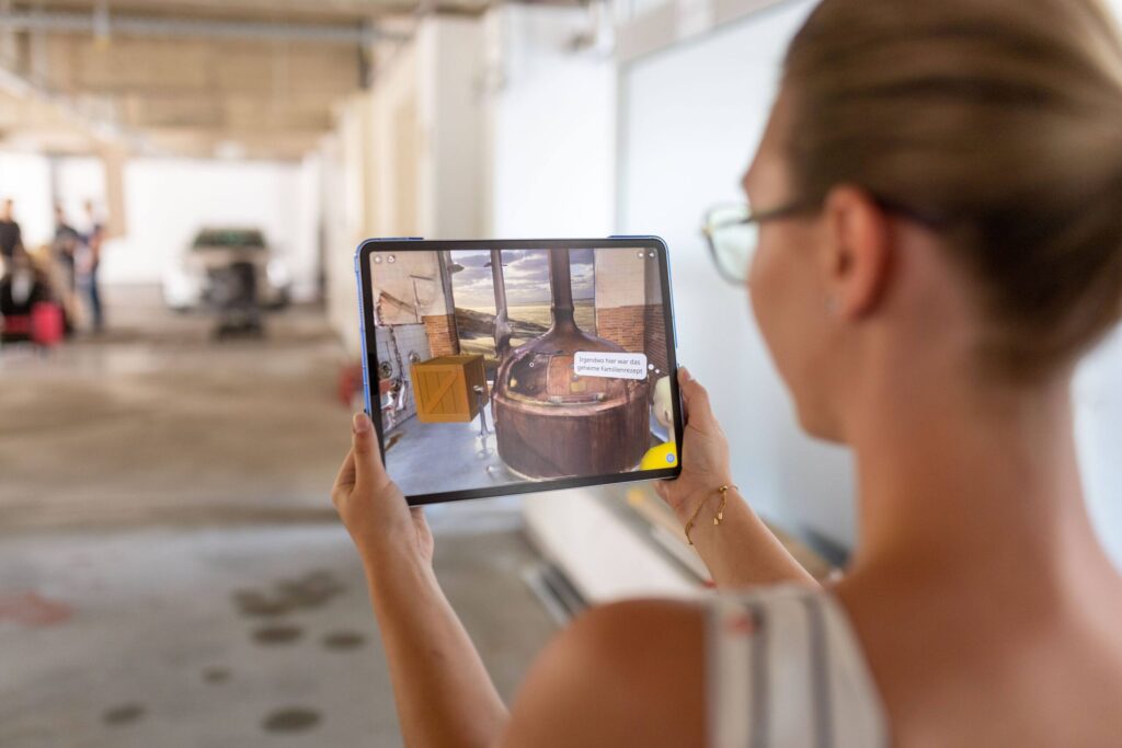 Eine Person hält ein Tablet mit Augmented-Reality-Ansicht eines Holzbottichs in einem geräumigen, hellen Innenraum – ideal, um innovative Anwendungen aus den Bereichen Wirtschaft oder Versicherungsmanagement zu erkunden. Die Person steht mit dem Rücken zur Kamera, trägt eine Brille und ein ärmelloses Oberteil. at Hochschule Coburg