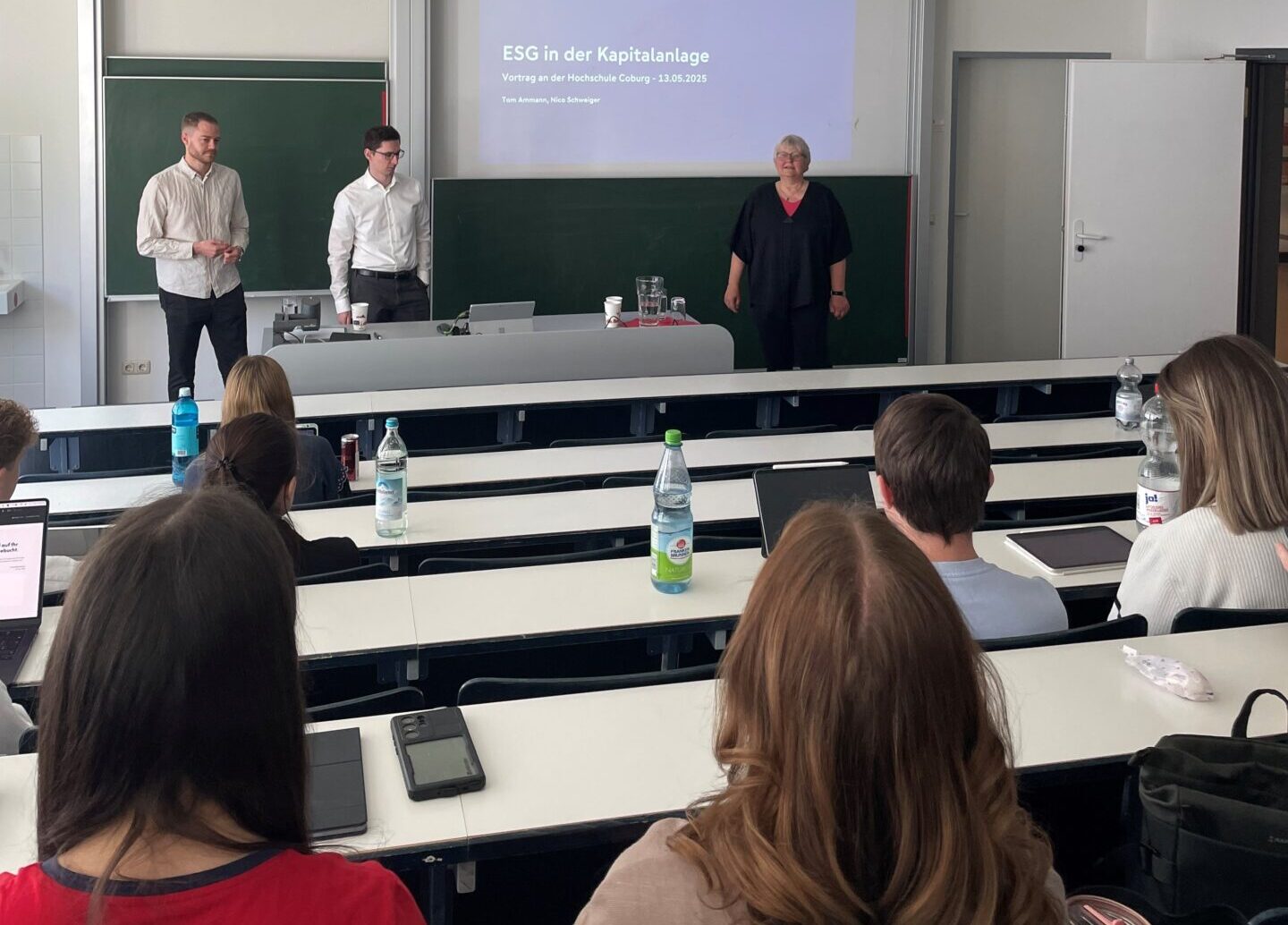 Drei Personen stehen vor einem Klassenraum und halten einen Vortrag mit dem Titel „ESG in der Kapitalanlage“. Sie diskutieren mit Schülern an ihren Tischen über Grüne Geldanlagen. Auf den Tischen stehen Wasserflaschen und Laptops. at Hochschule Coburg