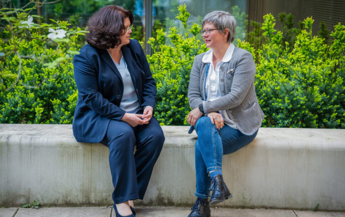 Zwei Frauen sitzen draußen auf einer Betonbank und unterhalten sich. Eine, vermutlich eine der Professorinnen, trägt einen marineblauen Anzug, die andere eine graue Jacke und Jeans. Grüne Büsche und ein Gebäudefenster bilden den Hintergrund. at Hochschule Coburg