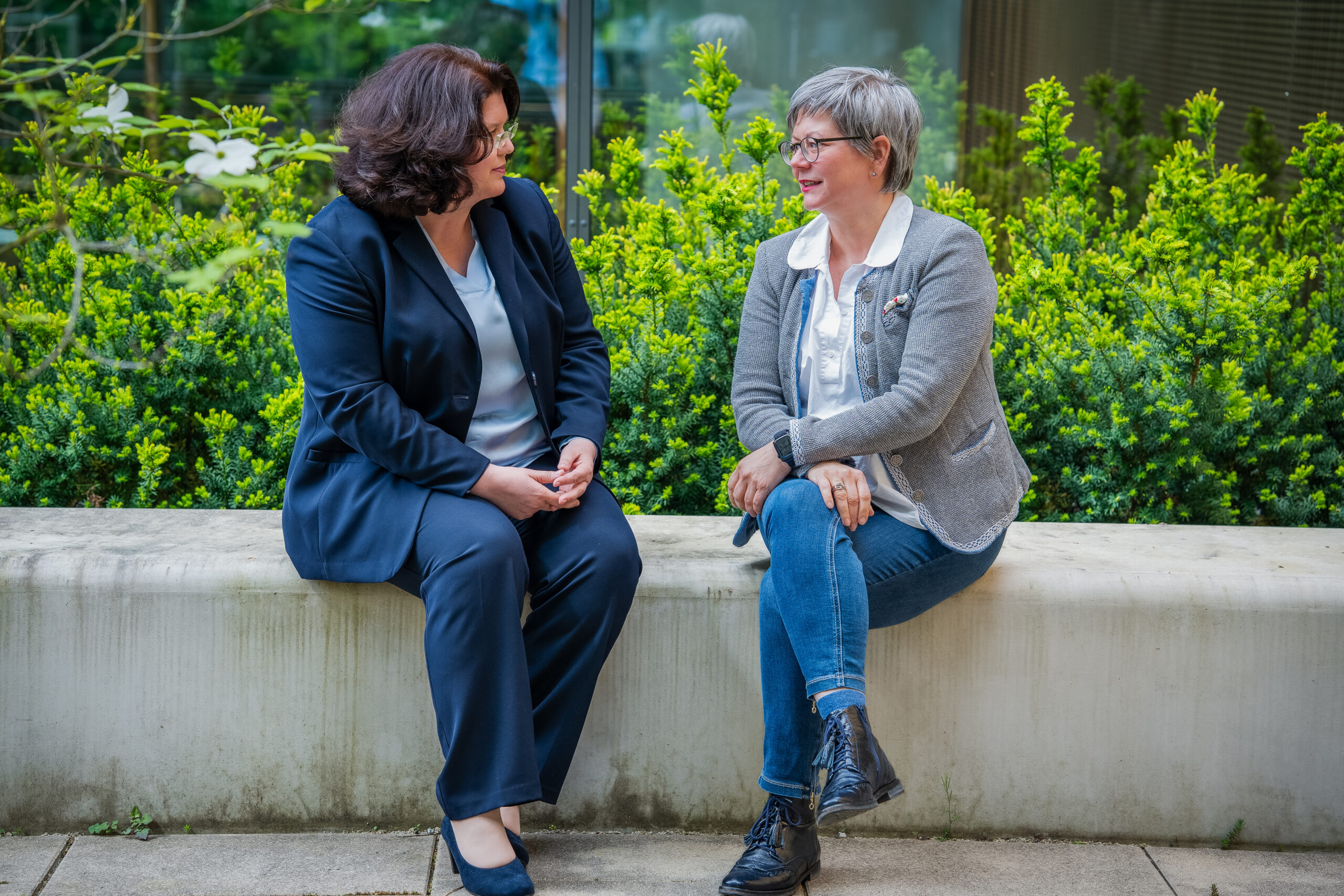 Zwei Frauen sitzen draußen auf einer Betonbank und unterhalten sich. Eine, vermutlich eine der Professorinnen, trägt einen marineblauen Anzug, die andere eine graue Jacke und Jeans. Grüne Büsche und ein Gebäudefenster bilden den Hintergrund. at Hochschule Coburg