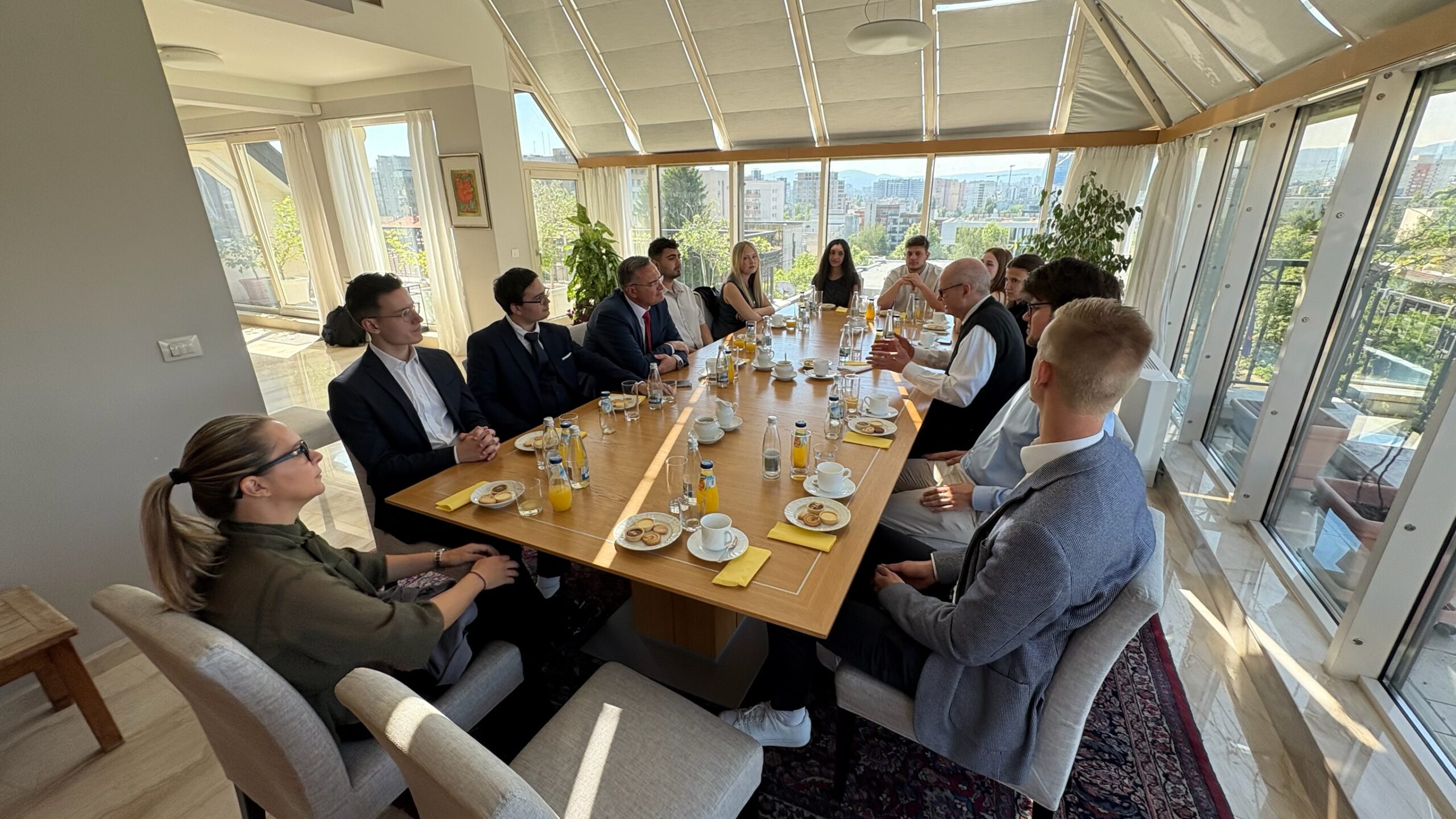 Eine Gruppe von Menschen in Geschäftskleidung sitzt zu einer Besprechung oder zum Essen um einen großen Tisch und diskutiert in dem hellen, sonnendurchfluteten Raum Wissen und Ideen – eine Szene aus einem deutsch-bulgarischen Studienprojekt mit Getränken und Tellern mit Essen auf dem Tisch. at Hochschule Coburg