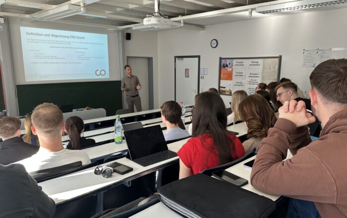 Eine Gruppe Studierender sitzt in einem hellen Hörsaal und hört einem Dozenten zu, der vorne im Raum Folien zu ESG-Scores und Grünen Geldanlagen präsentiert. Laptops und Notebooks stehen auf den Tischen. at Hochschule Coburg