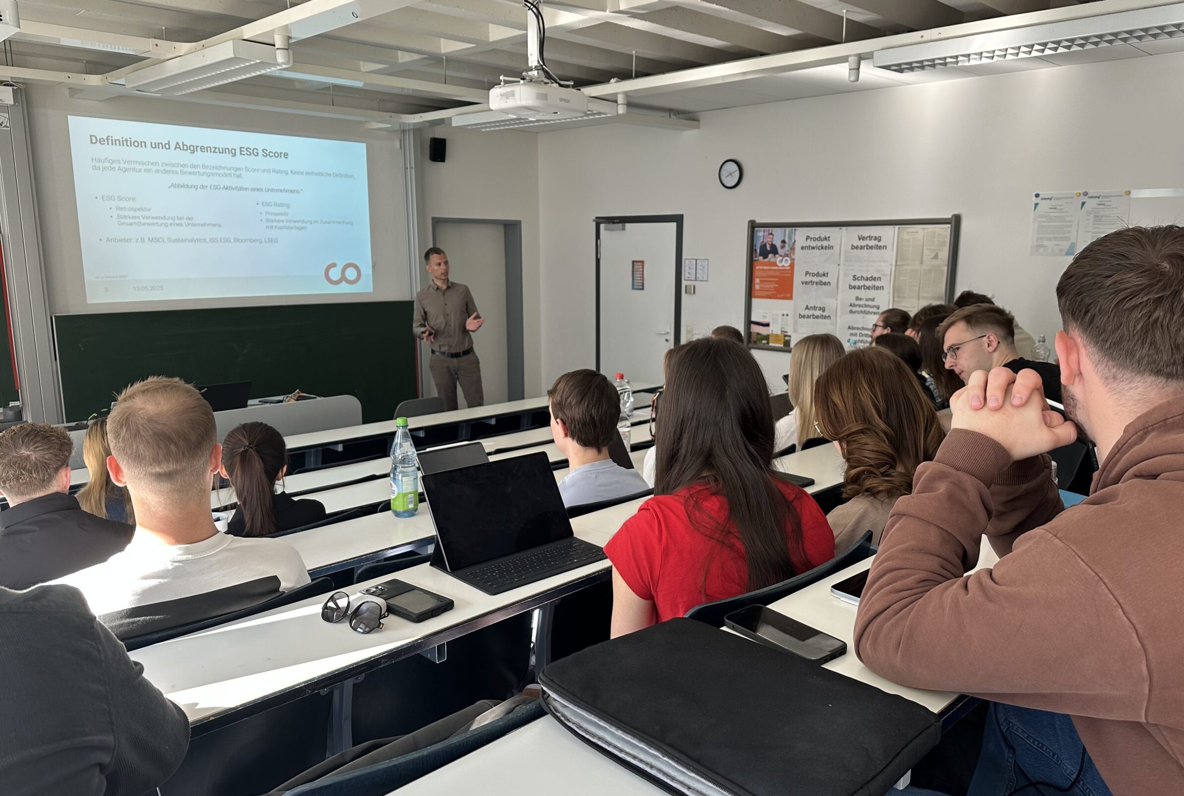 Eine Gruppe Studierender sitzt in einem hellen Hörsaal und hört einem Dozenten zu, der vorne im Raum Folien zu ESG-Scores und Grünen Geldanlagen präsentiert. Laptops und Notebooks stehen auf den Tischen. at Hochschule Coburg