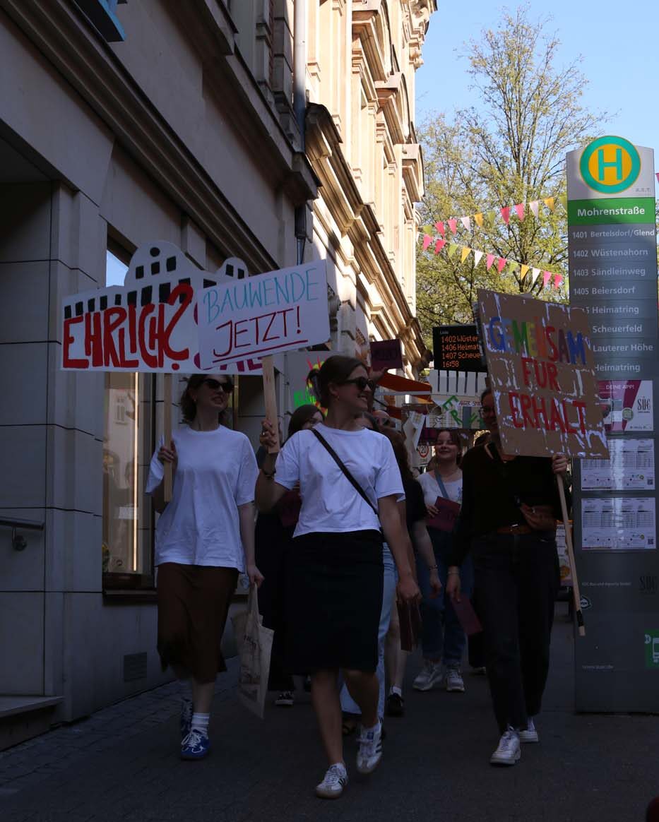 Eine Gruppe von Menschen, darunter Studierende, marschiert mit Protestschildern auf Deutsch auf einem Bürgersteig. Auf einem Schild steht „EHRLICH? BAUWENDE JETZT!“. Ein Bushaltestellenschild ist zu sehen, darüber hängen alte Gebäude und bunte Fahnen. at Hochschule Coburg